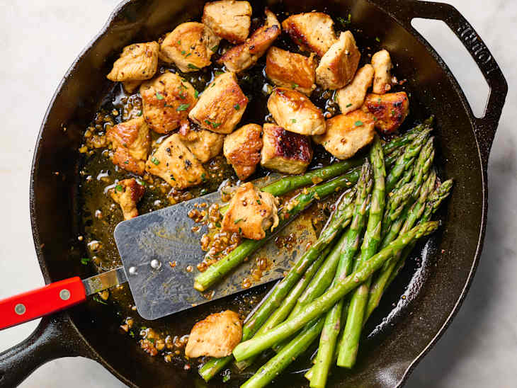 Sautéed chicken pieces with herbs and garlic alongside fresh asparagus in a cast iron skillet.