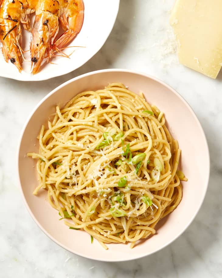 Overhead view of a garlic noodles in a pink bowl, a white bowl of prawns in the top left corner,and a corner of cheese.