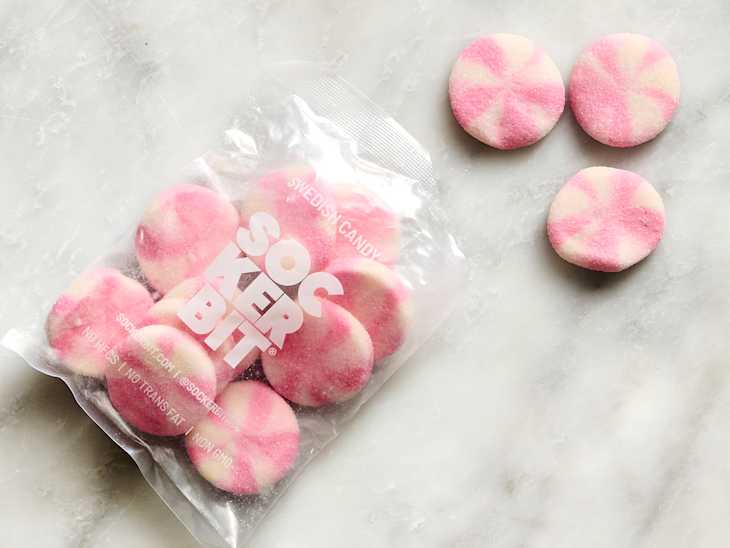 Colorful pink and white swirled Swedish candy pieces next to a clear bag on a marble surface.