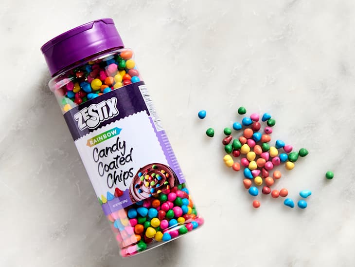 Container of rainbow candy-coated chips with colorful pieces scattered on a light surface.