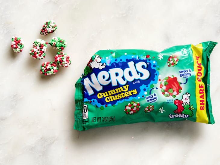 Colorful Nerds Gummy Clusters in festive red, green, and white, next to a green packaging pouch on a light surface.