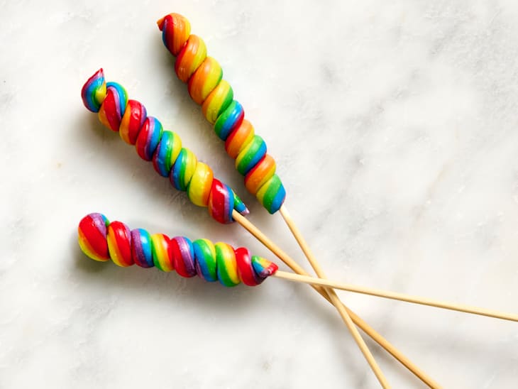 Colorful twisted candy skewers in red, yellow, green, and blue on wooden sticks against a light surface.