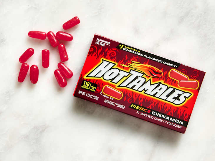 Red chewy candies scattered around a box of Hot Tamales Fierce Cinnamon flavored candy on a light surface.