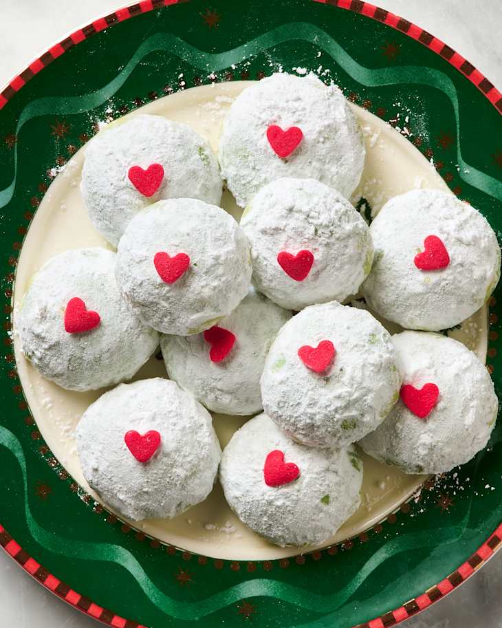 Snowball cookies dusted with powdered sugar and topped with red heart sprinkles on a festive plate.