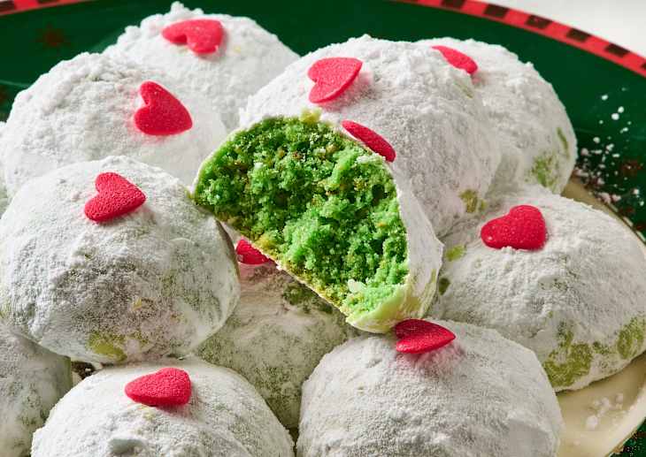 Green snowball cookies dusted with powdered sugar and topped with red heart sprinkles, one cookie cut in half.