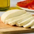 Sliced fresh cheese on a wooden cutting board with a plate of tomato slices and olive oil in the background.