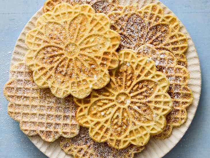 Decorative waffle cookies stacked on a textured plate, dusted with powdered sugar.