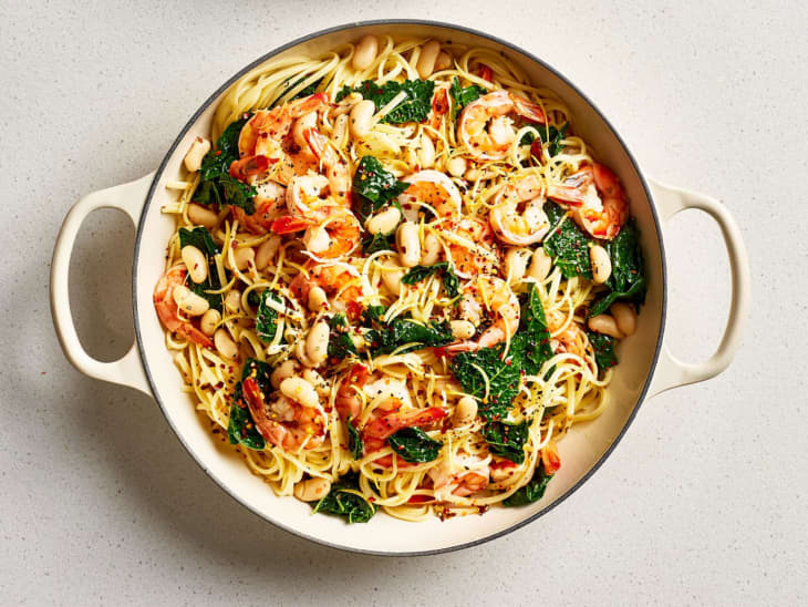 Shrimp pasta with kale and white beans in a large pot, garnished with lemon zest and pepper flakes.