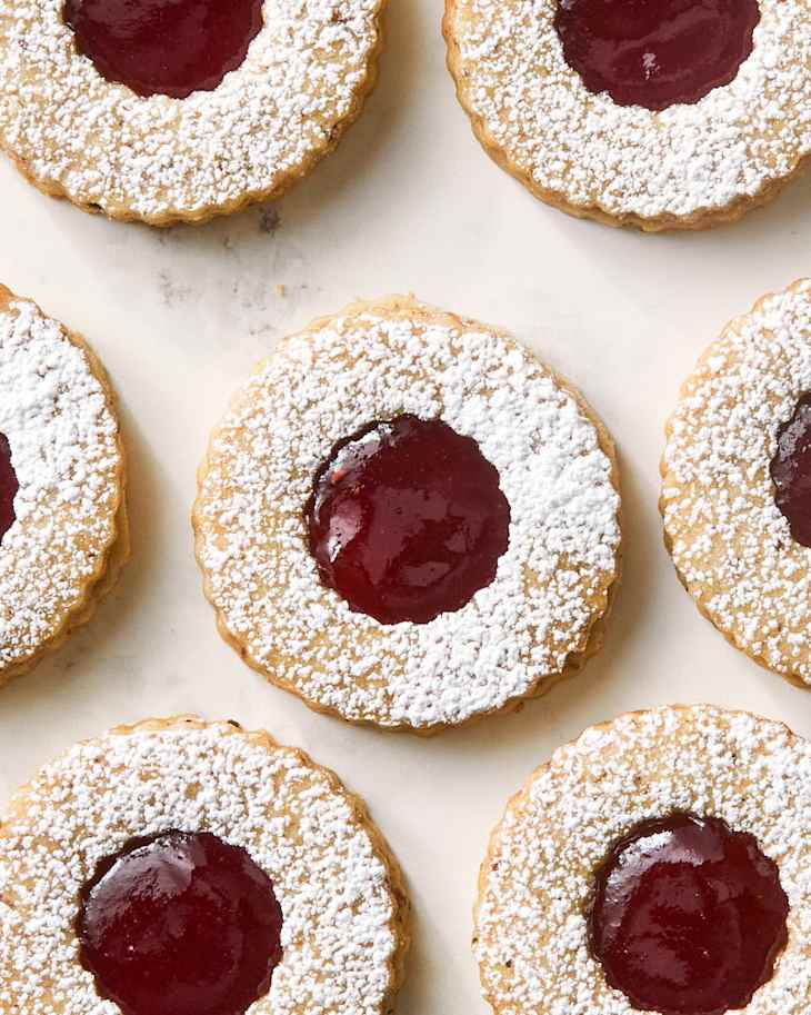Round cookies with a powdered sugar rim and a center of red jam, arranged on a light surface.