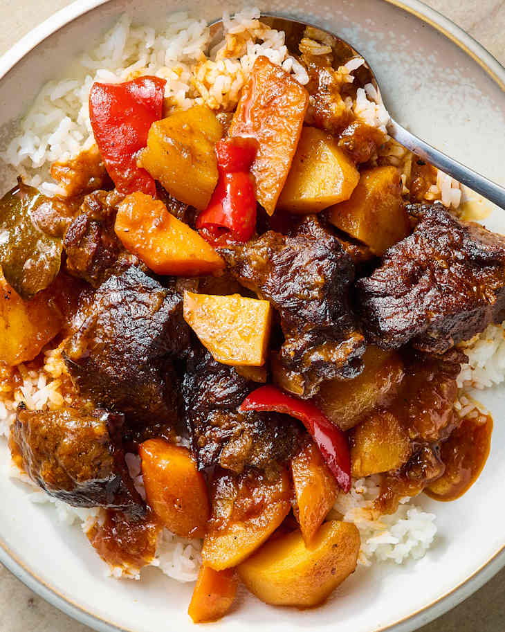 Savory beef stew with chunks of potatoes, red peppers, and rice in a white bowl.