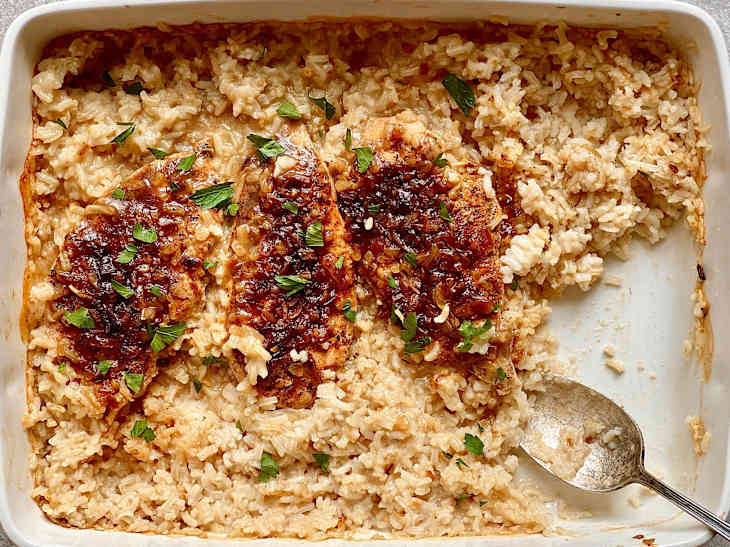 chicken baked in to cheesy rice in casserole dish with silver spoon portioned part out and green herbs on top