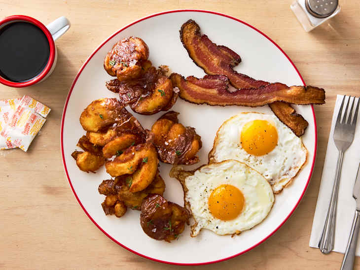 Breakfast plate featuring crispy bacon, two sunny-side-up eggs, and golden-brown potato pieces, with a cup of coffee.