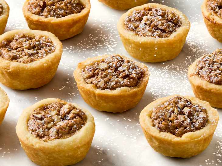 Mini pecan pies with a golden crust, topped with powdered sugar, arranged on a baking sheet.