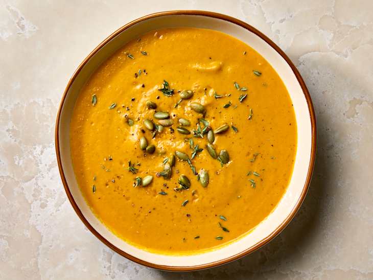 Creamy orange soup topped with pumpkin seeds and fresh thyme in a shallow bowl.