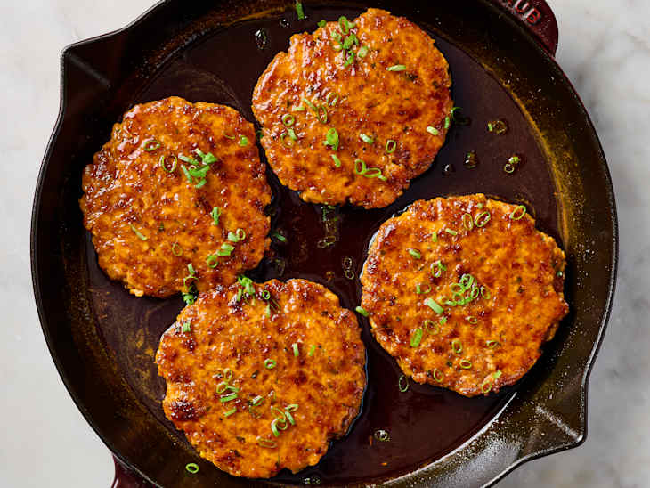 Four salmon cakes garnished with chopped green onions in a dark skillet.