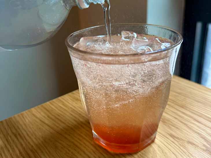 Someone making a shrub drink by pouring seltzer into a glass with ice and Kosterina Crushed Strawberry Vinegar