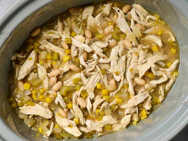 Shredded chicken, white beans, corn, and green chilies in a slow cooker with broth and diced onions.