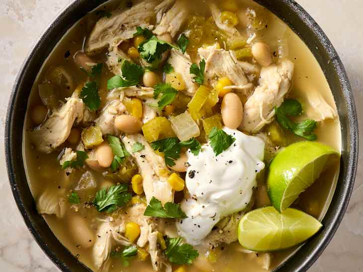 Bowl of chicken soup with shredded chicken, white beans, corn, green chilies, cilantro, and a dollop of sour cream, garnished with lime.