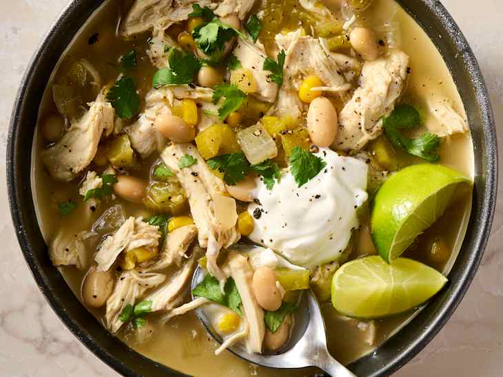 Bowl of chicken soup with shredded chicken, white beans, corn, green chilies, cilantro, and a dollop of sour cream, garnished with lime.