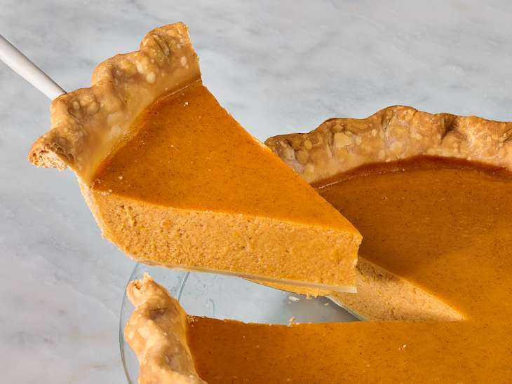 a slice of sweet potato pie being lifted from a whole pie with a flaky crust, set on a light marble surface.