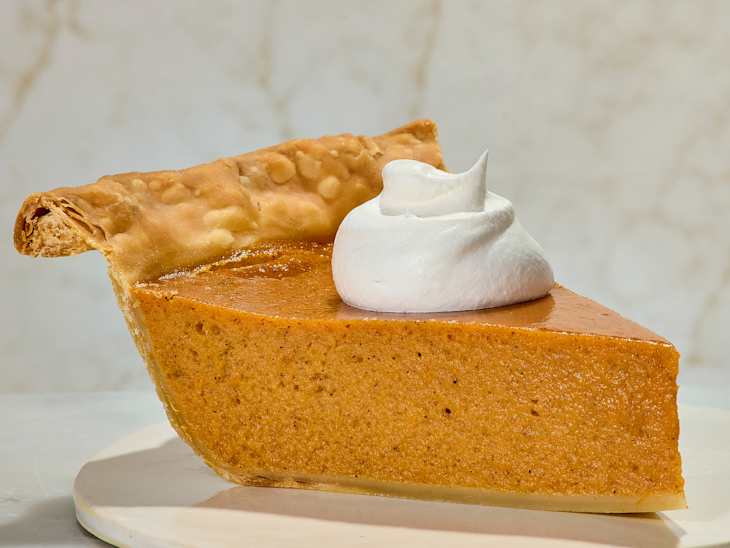 Slice of sweet potato pie with a flaky crust, topped with a swirl of whipped cream on a white plate.